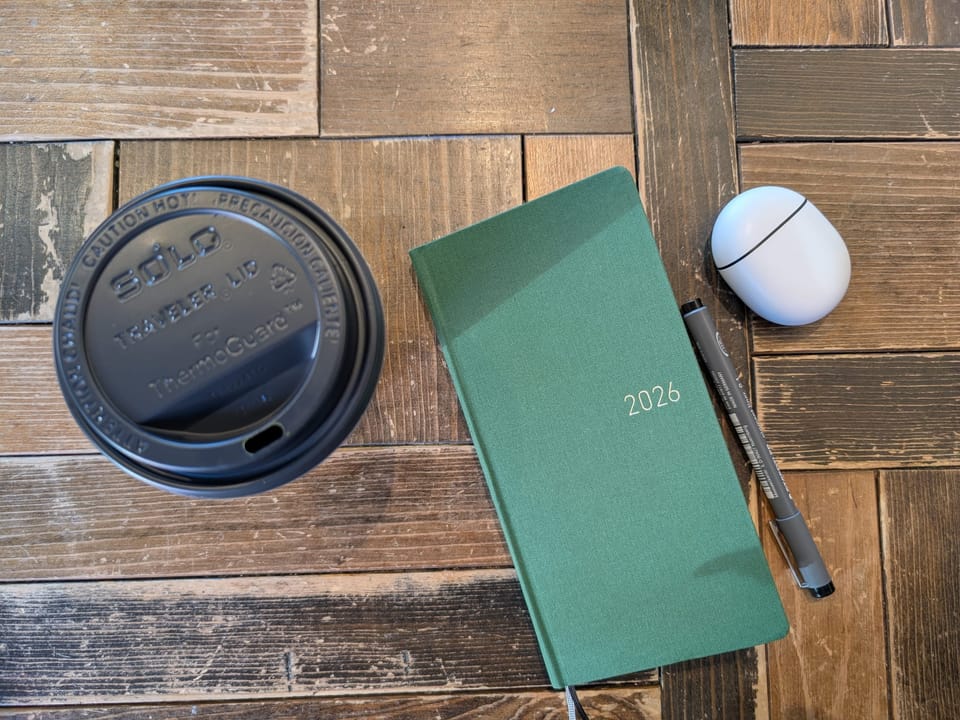 Looking down on a table with a green Hobinichi Weeks, a grey pen, a headphone case, and a to go cup from a coffee shop.